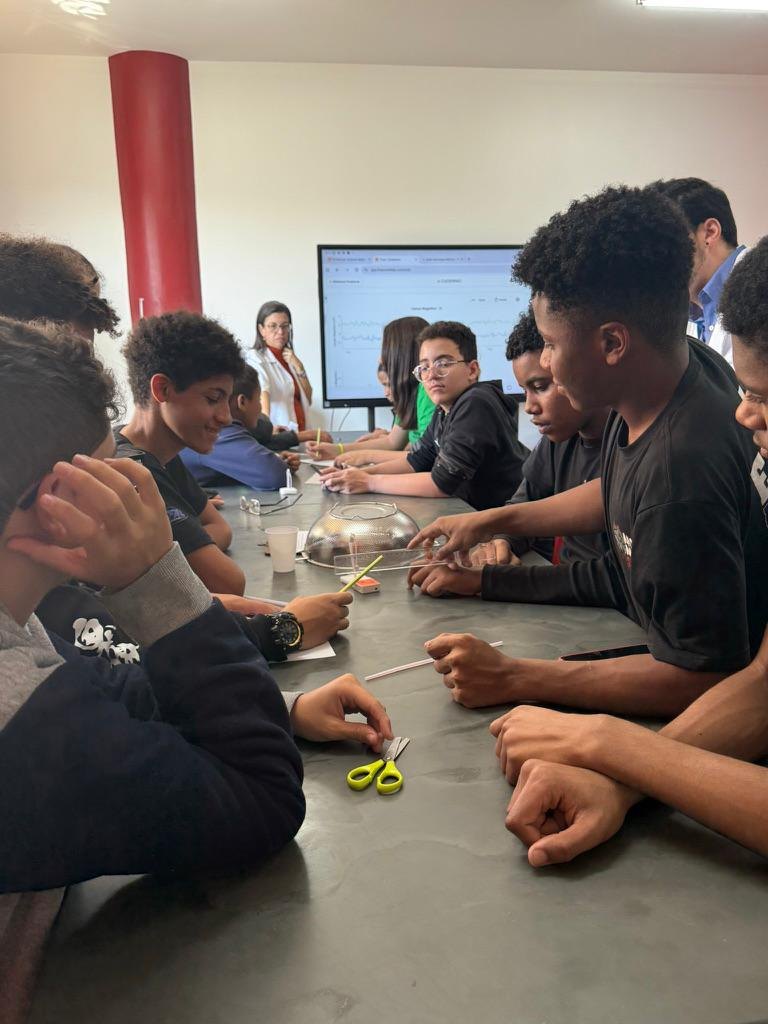 Alunos ao redor de uma mesa durante o experimento da gaiola de Faraday, com uma peneira de metal sobre a mesa