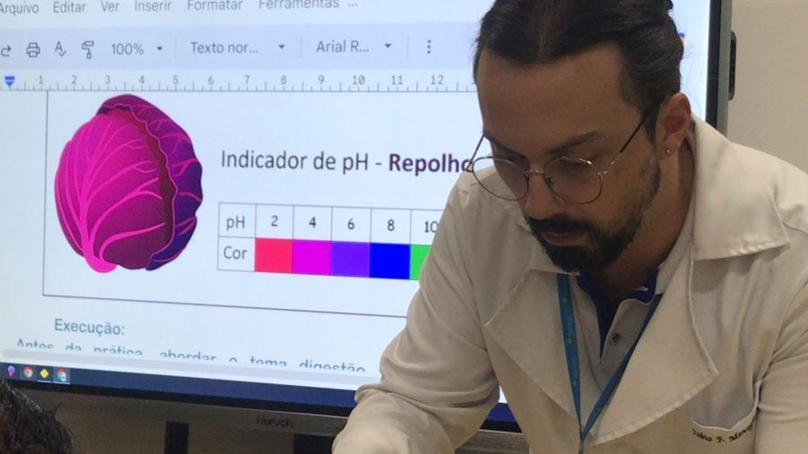 Professor Fábio Fialho Meneghesso em aula de Química, com a tabela de cores do indicador de pH de repolho roxo projetada na tela