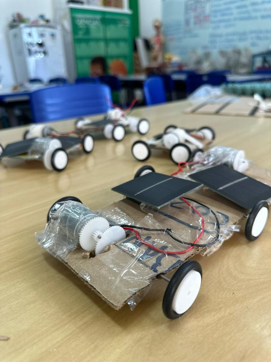 Carrinho de papelão com motor, engrenagens brancas, rodas e dois painéis solares, sobre uma mesa de sala de aula